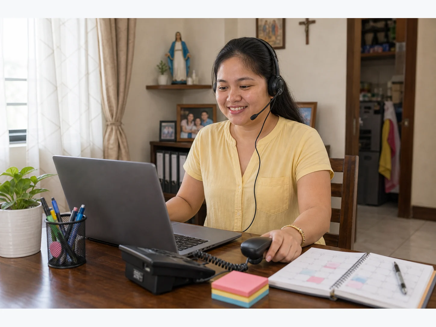 Filipina medical receptionist in a yellow blouse working from a home dining-table workspace with a laptop, phone, headset, and appointment notes.